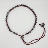 A beaded necklace with small dark gray and black marbled beads threaded on a deep red cord, featuring a round silver-toned medallion and adjustable sliding cord ends, displayed on a clean white background.