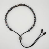 A beaded necklace made of small dark, iridescent oval beads on a black cord, with a round silver-toned central disc and adjustable cord ends, photographed on a plain light background.